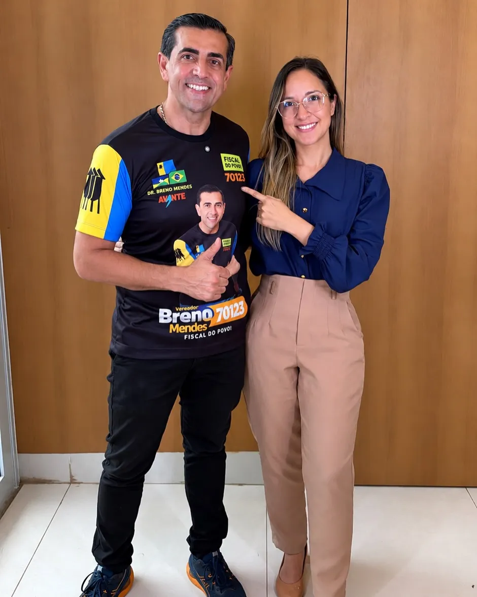 Dr. Breno Mendes de camisa da campanha faz sinal de positivo ao lado de uma apoiadora de blusa azul que aponta para ele, em frente a uma parede de madeira.
